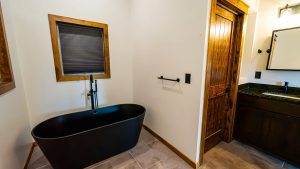 Black freestanding bathtub with a matte finish sits in a modern bathroom, framed by wooden trim and a gray tiled floor.