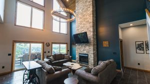 Spacious living room with a circular chandelier, tall stone fireplace, and a mounted TV above the fireplace; large windows and a sliding door to a deck overlook a suburban neighborhood.