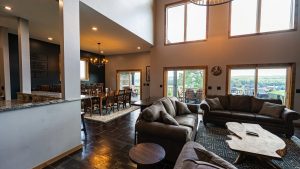 Open-concept living area with brown couches, a stone coffee table, and large windows overlooking greenery outside.