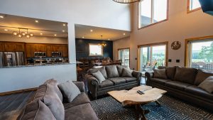 Open-concept living area with gray sectional sofas, a rustic coffee table, a kitchen on the left, dining area in the back, and large windows showing a hillside view.