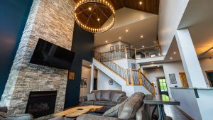 Living room with a stone fireplace, a large circular chandelier, and a wooden staircase railing overlooking the lower level.
