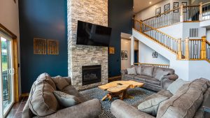 Open-concept living room with a stone fireplace, mounted TV, and dark blue accent wall behind beige sectional sofas.