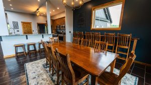 Open-plan kitchen and dining area with a long wooden table and matching chairs, a dark blue accent wall, and a large window.