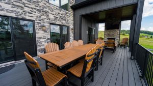 Outdoor deck dining area with a long wooden table and Adirondack chairs beside a stone building and large windows, overlooking rolling hills.