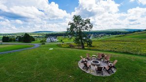 Circular stone fire pit surrounded by wooden Adirondack chairs in a grassy backyard, with a row of houses and green hills in the distance under a cloudy sky.