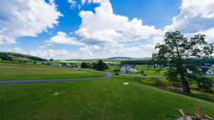 Wide view of a suburban landscape with green lawns, a curved road, and scattered houses beneath a bright blue sky filled with white clouds, and a large tree on the right.