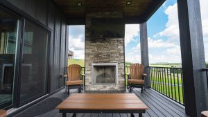 Outdoor patio with a stone fireplace and wall-mounted TV, two brown Adirondack chairs, and a wooden table, overlooking a green landscape.