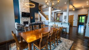 Open living and dining area featuring a long wooden table, stone fireplace with TV, blue accent wall, and a staircase with wooden railings.