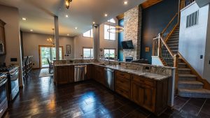 Open-concept kitchen and living area with island, granite countertops, stainless appliances, and a stone fireplace near a wooden staircase.