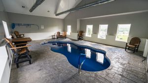 Indoor pool area with a blue, irregularly shaped pool, gray walls, exposed ductwork, and wooden Adirondack chairs along the walls.