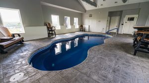 Indoor pool area with a curved blue pool, gray tiled floor, and wooden chairs along a windowed wall.