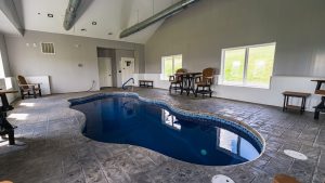 Indoor pool area with gray walls, large windows, and wooden chairs around high tables—blue water visible in a curved pool edge.