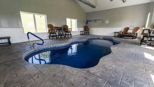 Indoor pool area with a curved blue pool, gray tile floor, and wooden lounge chairs along the walls in a bright room.
