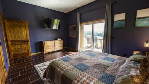 Bedroom with navy walls, wooden armoire and dresser, and a bed facing a balcony door opening to a hillside view.