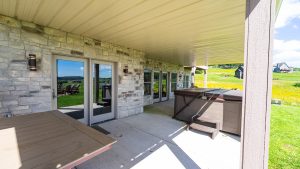 Covered patio attached to a stone house with glass doors; a large table sits on the concrete deck, overlooking a green, rural landscape.
