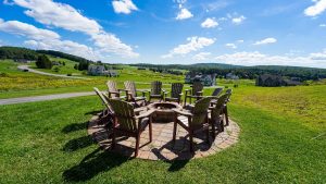 Adirondack chairs circle a fire pit on a brick patio, overlooking green lawn and houses under a bright blue sky.