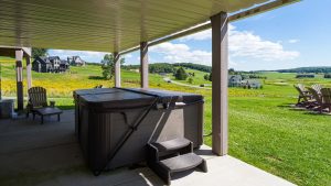 Covered patio with a large hot tub on a concrete slab, steps to the yard, and yellow wildflowers in the distance under a blue sky with white clouds; Adirondack chairs visible to the right.