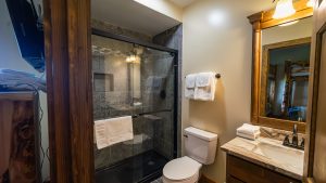 Bathroom interior with a glass-enclosed shower, gray tile, white towels, and a wooden vanity with a large mirror in a rustic cabin-style room.