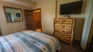 Rustic bedroom with a quilted blue bedspread, a large log-style dresser under a wall-mounted TV, and a wide mirror on the adjacent wall.