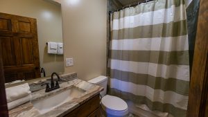Bathroom with a marble countertop double vanity, black faucets, towel rack, toilet, wooden door, and beige-white striped shower curtain.