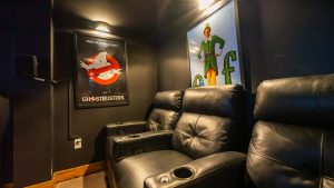 Dim home theater with black leather recliners; Ghostbusters poster on the left wall and Elf poster on the right wall.