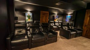 Dim home theater with rows of black leather reclining chairs and movie posters on dark walls.