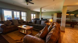 Spacious living room with brown sectional sofas, patterned throw pillows, and a wooden coffee table by large windows exposing outdoors. A pool table is visible in the back.
