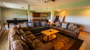 Open-concept living area with a brown leather sectional, patterned throw pillows, and a wooden coffee table, pool table visible in the back.