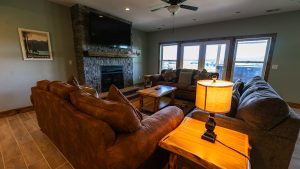 Cozy living room with a stone fireplace, mounted TV, and brown sectional sofa facing large windows that overlook an outdoor area.