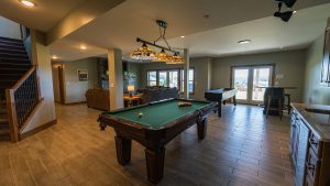 Spacious basement game room with a central pool table, warm hanging lamps, and a seating area by a stone fireplace.