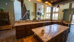 Open-concept kitchen with marble countertops, dark wood cabinets, and a stone fireplace visible in the living room; staircase to the left.