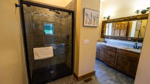 Modern bathroom with a glass shower enclosure, dark stone tiles, and a wooden vanity reflected in a large framed mirror.