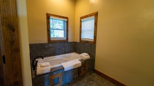 Corner bathroom with a white tub set in dark stone surround, two small windows with white blinds, and warm yellow walls.