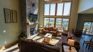 Cozy living room with a stone fireplace, mounted TV, and large windows that overlook a sunny outdoor deck and green fields; brown sofas and wooden coffee tables dominate the seating area.