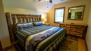 Rustic log-bedroom with a large wooden bed, plaid quilt, and matching dresser and mirror by a window.
