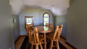 Round wooden dining table with log chairs in a small sunlit dining nook with a sloped ceiling and arched window.