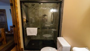 Glass-enclosed shower with dark marble tiles, a white towel on a rack, and a nearby white toilet in a wood-framed bathroom.