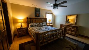Rustic log-bedroom with a large wooden bed, matching nightstands, lamps, and a dresser with a framed mirror; cabin decor and a pillow that reads 'CABIN IS MY HAPPY PLACE'.