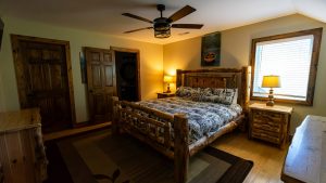Rustic cabin bedroom with a log-frame bed, matching nightstands, and two lamps; window with blinds on the right.