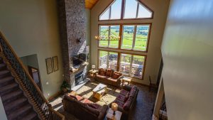 Two-story living room with a stone fireplace, large windows, and a chandelier overlooking a green landscape.