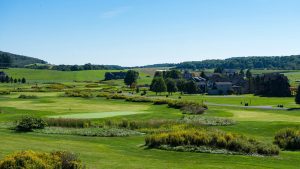 Wide view of a green golf course with fairways, shrubs, and houses in the distance under a clear blue sky.