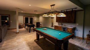 Basement game room with a green-felt pool table, three stained-glass pendant lights, and a wooden bar area with cabinets in the back.