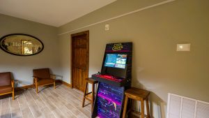 Upright arcade racing game cabinet in a cozy lounge with tan walls, wooden doorway, and brown chairs nearby.