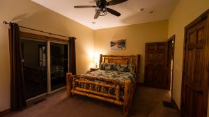 Rustic bedroom with a heavy log-frame bed, patterned bedding, a bedside lamp, and a sliding glass door with dark curtains.