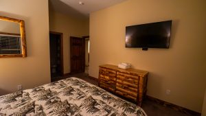 Hotel room with a mountain-pattern bedspread, a rustic log dresser, and a wall-mounted flat-screen TV on a beige wall; doorway to a bathroom is open in the background.