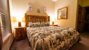 Cozy cabin bedroom with a log bed, matching nightstands, warm lamps, and forest-themed bedding plus a wooden-framed mirror on the wall.