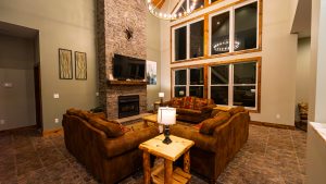 Cozy living room with a stone fireplace, antler mount, and large wood-trimmed windows; brown leather sofas surround a rustic wooden coffee table.