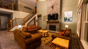 Cozy living room with brown leather sofas, wooden coffee table, stone fireplace, and staircase with iron railing.