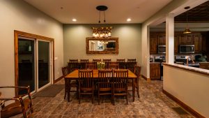 Dining room with a long wooden table and many chairs, warm pendant lights, and a rustic framed mirror on a green wall over the table.