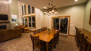 Rustic dining room with a long wooden table and matching chairs, a multi-lantern chandelier overhead, and a stone fireplace on the left side.